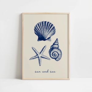 Puede incluir: Una impresión enmarcada con ilustraciones azules de una concha, una estrella de mar y una concha en espiral sobre un fondo crema. Las palabras "sun and sea" están escritas en azul. El marco es de madera clara.