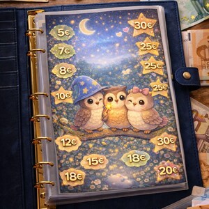 Puede incluir: Un archivador azul con detalles dorados, abierto para revelar un rastreador de ahorro con una ilustración caprichosa de tres búhos en una rama bajo un cielo estrellado. El rastreador tiene cantidades en euros.