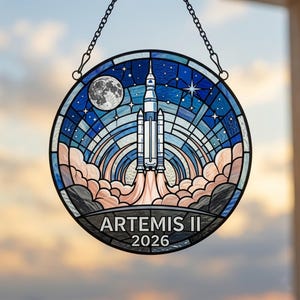 May include: A round stained glass panel depicting a rocket launching into space, with a full moon and stars in a blue sky. The words "ARTEMIS II 2026" are at the bottom. The panel is suspended by a chain.