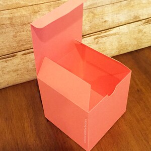 May include: A coral pink, square-shaped gift box with a hinged lid. The box is open and the inside is visible. The box is made of sturdy cardboard and has a smooth finish. The text "amnieslittleboxes" is printed on the side of the box.