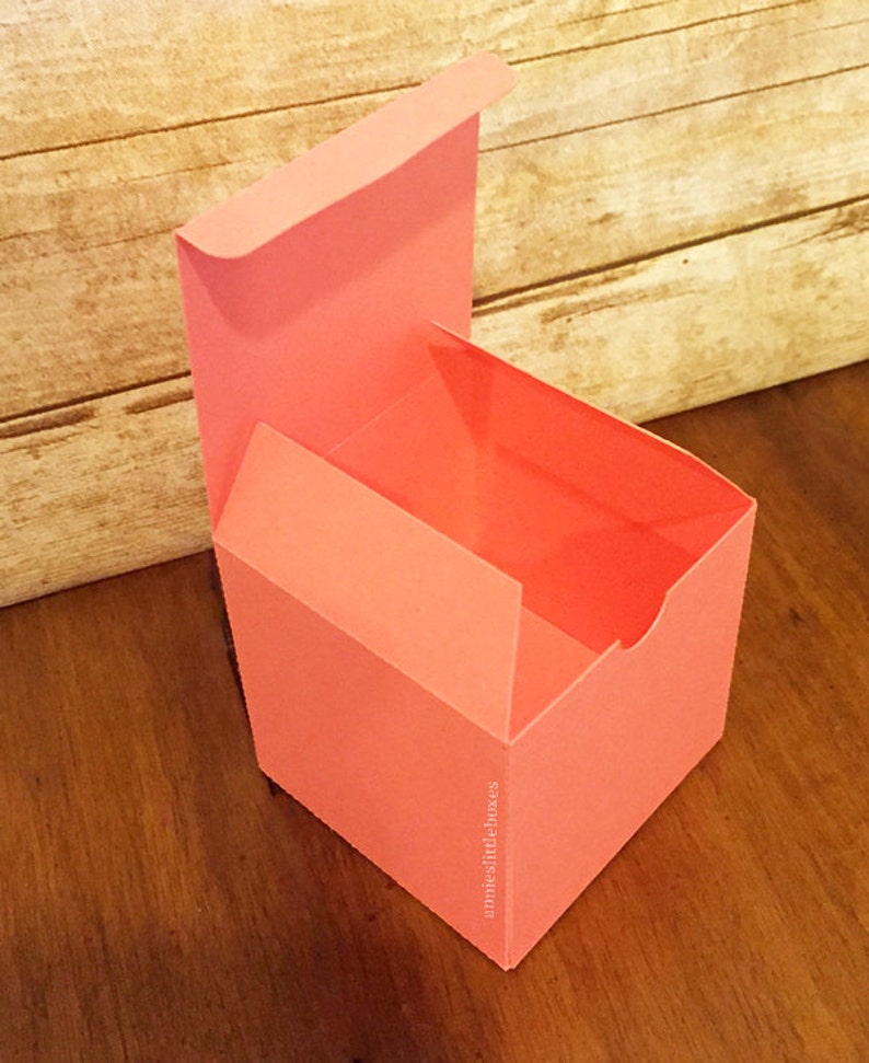 May include: A coral pink, square-shaped gift box with a hinged lid. The box is open and the inside is visible. The box is made of sturdy cardboard and has a smooth finish. The text "amnieslittleboxes" is printed on the side of the box.