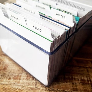 Pode incluir: Uma caixa de armazenamento de plástico transparente com divisores etiquetados com várias categorias, incluindo "Primavera", "Natal", "Aniversário", "Criaturas", "Olá", "Inverno", "Halloween" e "Floral".