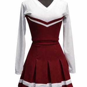 Op de afbeelding: Een bordeauxrood en wit cheerleaderuniform op een mannequin. De top is wit met een bordeauxrood chevron-ontwerp en lange mouwen. De geplooide rok is bordeauxrood met witte strepen aan de zoom. Een klassieke outfit voor cheerleading.