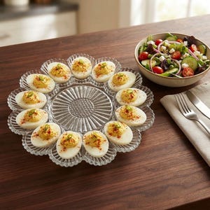 May include: A clear glass deviled egg platter with a scalloped edge and a central sunburst pattern, holding twelve halved hard-boiled eggs topped with yellow yolk filling, red paprika, and green onion slices, arranged in a circular formation.