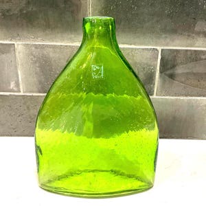 May include: A large, green glass bottle with a wide base and narrow neck. The bottle has a textured surface and a small logo that reads "AMICI". The bottle is empty and sits on a white surface.