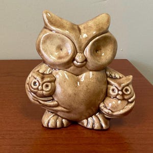 May include: A ceramic owl figurine in shades of brown, holding two smaller owl figures. The owl has large, round eyes and a smiling expression. The figurine is a decorative item, likely for a shelf or tabletop.