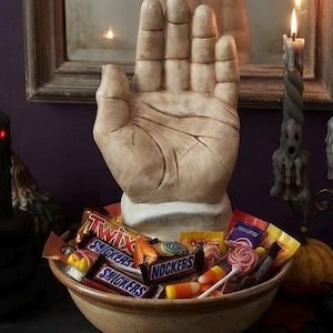 May include: A beige ceramic bowl filled with assorted candy bars including Twix, Snickers, and Nockers, topped with a large, upright, light-colored sculpted hand figurine. The hand features visible palm lines and sits centered in the bowl among the candy.