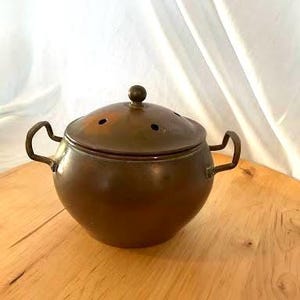 May include: A round, metallic, copper-toned pot with two side handles and a matching domed lid. The lid features a small central knob and several circular ventilation holes. The pot has a smooth, curved body and a warm, burnished finish.