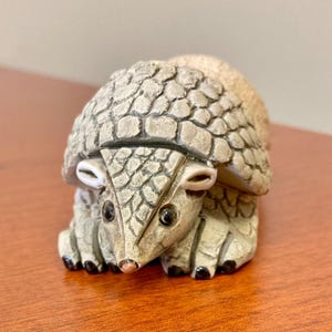 May include: A detailed figurine of an armadillo, featuring a textured, segmented shell in shades of gray and tan. The armadillo has small, dark eyes, a pink nose, and black claws. The figurine is posed in a crouching position.