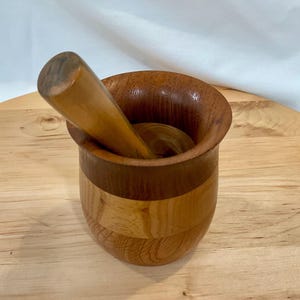 May include: A wooden mortar and pestle set featuring a two-tone design. The mortar is constructed from horizontal bands of light and dark wood, while the pestle is a smooth, solid piece of light-colored wood resting inside the bowl.