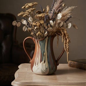 May include: A ceramic pitcher with two handles featuring a brown base and a white and blue drip glaze finish. The pitcher is filled with a dried floral arrangement consisting of tan yarrow, purple lavender, pampas grass, and silver dollar plant stems.