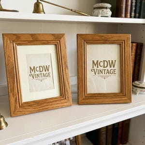 May include: Two rectangular wooden picture frames with a light brown finish. Each frame has a cream-colored mat and the text "MCDW VINTAGE" in a decorative font. The frames are displayed on a white shelf.