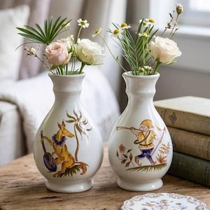 May include: Two white ceramic vases with narrow necks and rounded bodies. The left vase features a golden fox playing a stringed instrument, while the right vase depicts a small figure in a yellow tunic blowing a horn. Both contain fresh flowers.