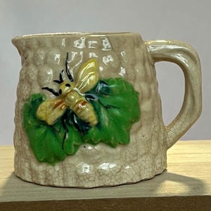 May include: A small, beige ceramic pitcher with a handle. The pitcher is decorated with a raised relief of a yellow and black bee on a green leaf. The pitcher has a textured surface and a crackled glaze.