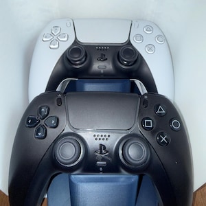 Op de afbeelding: Een wit en zwart draadloze gamecontroller boven een volledig zwarte draadloze gamecontroller, beide rustend op een donkerblauw plastic oplaadstation. De controllers zijn voorzien van twee analoge sticks, een D-pad en vier actieknoppen.