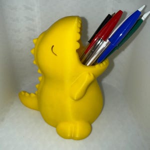 May include: A bright yellow dinosaur-shaped pencil holder with a wide-open mouth containing several pens. The dinosaur has a smooth, matte finish, a curved tail, and small spikes along its back, serving as a functional desk organizer for pens.