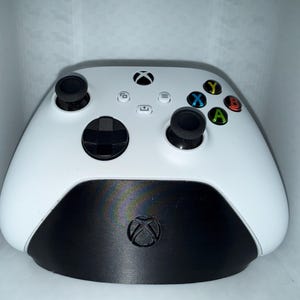 May include: A white Xbox controller with black accents. The controller features black joysticks, a directional pad, and colored action buttons. The Xbox logo is visible on the controller and the base.