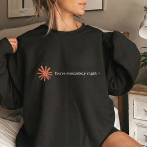 Puede incluir: Una sudadera negra de manga larga con un pequeño gráfico de rayos de sol en color terracota en el pecho izquierdo. Junto al gráfico, el texto en blanco dice "You're absolutely right -" en una fuente sencilla, clara y sin remates.