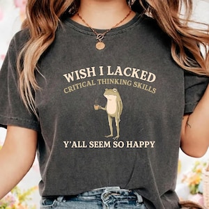 Puede incluir: Una camiseta de manga corta gris carbón con el gráfico de una rana de pie sosteniendo una taza pequeña. El texto dice WISH I LACKED CRITICAL THINKING SKILLS Y'ALL SEEM SO HAPPY en un color crema claro. La camiseta tiene cuello redondo.