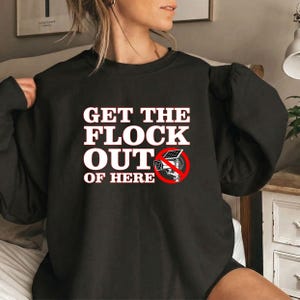 May include: A black long-sleeve sweatshirt featuring the white text "GET THE FLOCK OUT OF HERE" and a graphic of a security camera with a solar panel, enclosed within a red circular prohibition symbol with a diagonal line through it.