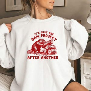 May include: A white long-sleeve sweatshirt featuring a red graphic of a beaver gnawing on a log next to a stack of wood. The text printed in red reads, IT'S JUST ONE DAM PROJECT AFTER ANOTHER, positioned above and below the beaver illustration.
