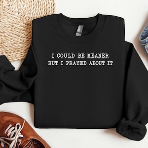 Könnte beinhalten: Ein schwarzes langärmeliges Sweatshirt mit Rundhalsausschnitt und dem weißen Text "I COULD BE MEANER BUT I PRAYED ABOUT IT", der mittig auf der Brust aufgedruckt ist. Das Kleidungsstück hat einen gerippten Ausschnitt, Bündchen und Saum.