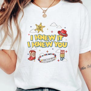 May include: A white t-shirt featuring a graphic print with the text I KNEW IT I KNEW YOU in yellow. The design includes a gold sheriff star, a red cowboy hat, red cowboy boots, a beaded bracelet with the text YOU'VE GOT A FRIEND IN ME, and blocks.
