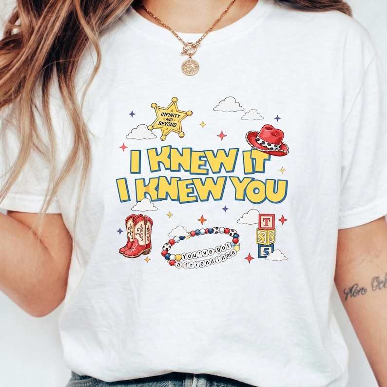 May include: A white t-shirt featuring a graphic print with the text I KNEW IT I KNEW YOU in yellow. The design includes a gold sheriff star, a red cowboy hat, red cowboy boots, a beaded bracelet with the text YOU'VE GOT A FRIEND IN ME, and blocks.