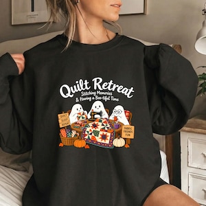 May include: A black crewneck sweatshirt featuring a graphic of three white ghosts quilting at a table. The text reads Quilt Retreat Stitching Memories & Having a Boo-tiful Time, with signs saying Quilt Sleep Repeat and Fabric Friends Fun.