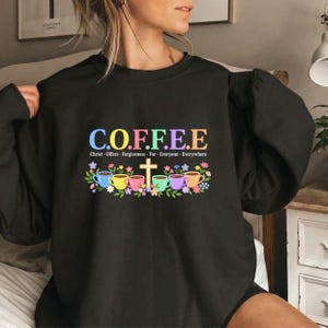 May include: A black long-sleeve sweatshirt featuring the colorful text C.O.F.F.E.E. with the acronym Christ Offers Forgiveness For Everyone Everywhere. Below the text is a graphic of five colorful coffee mugs, a gold cross, and flowers.
