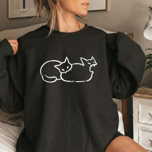 Könnte beinhalten: Ein schwarzes Langarm-Sweatshirt mit einer weißen Strichgrafik von zwei Katzen, die nebeneinander positioniert sind. Das Design zeigt eine zusammengerollte Katze und eine Katze mit geöffnetem Maul in einem einfachen, weißen Umrissstil.