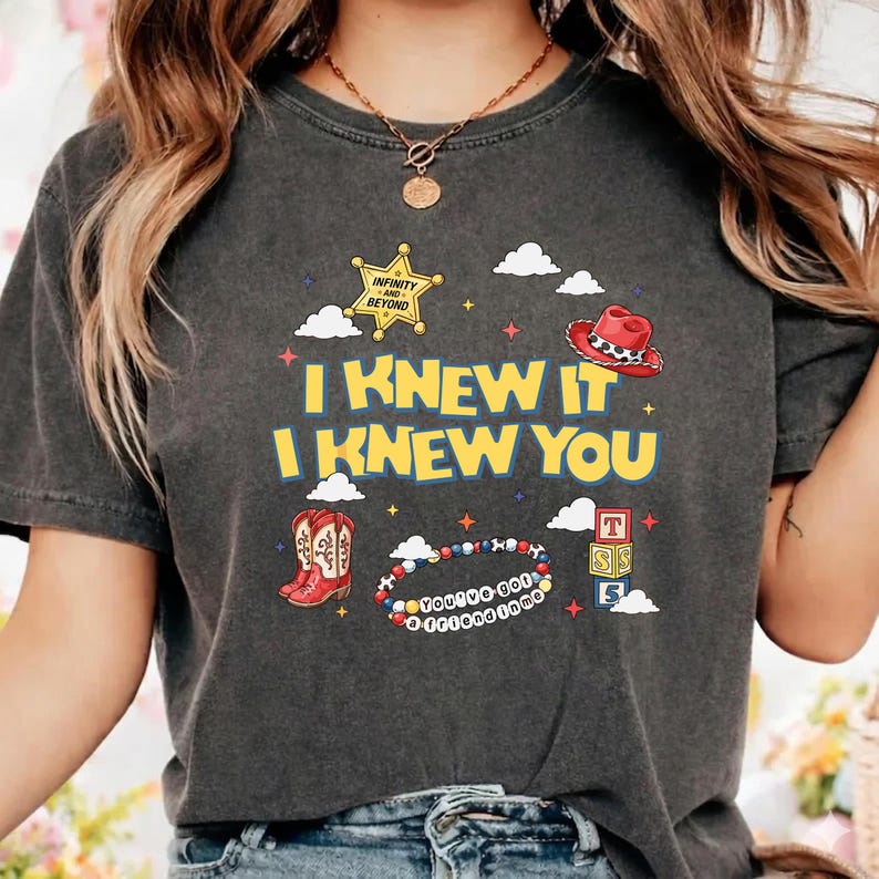 May include: A charcoal gray t-shirt featuring a graphic print with the yellow text I KNEW IT I KNEW YOU. The design includes a sheriff badge, a red cowboy hat, red cowboy boots, a beaded bracelet with text, alphabet blocks, and white clouds.