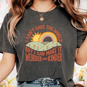 May include: A charcoal gray short-sleeve t-shirt featuring a graphic print of a yellow sun, a rainbow, and green hills with small flowers. The text reads: I CAN'T SAVE THE WORLD BUT I CAN MAKE IT WEIRDER AND KINDER in orange and red lettering.