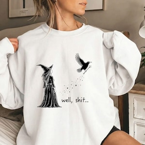 May include: A white crewneck sweatshirt featuring a black graphic print of a witch in a pointed hat and long dress standing near a flying crow holding a star-tipped wand. The text "well, shit.." is printed in black below the crow.