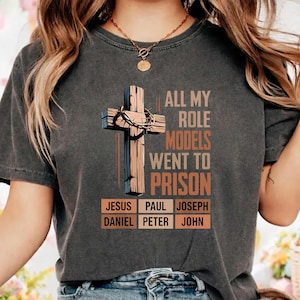 May include: A charcoal gray short-sleeve t-shirt featuring a graphic of a wooden cross with a crown of thorns. The text reads: ALL MY ROLE MODELS WENT TO PRISON, followed by a grid of names: JESUS, PAUL, JOSEPH, DANIEL, PETER, and JOHN.