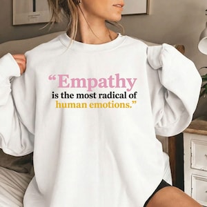 Może przedstawiać: Biała bluza z długim rękawem z napisem "Empathy is the most radical of human emotions." Słowo "Empathy" jest nadrukowane na różowo, "is the most radical of" na czarno, a "human emotions." na żółto, wszystko czcionką szeryfową.