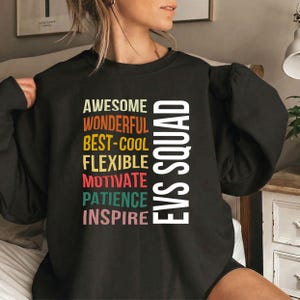 May include: A black crewneck sweatshirt featuring the text EVS SQUAD in large white letters. To the left, a list of words appears in various colors: AWESOME, WONDERFUL, BEST-COOL, FLEXIBLE, MOTIVATE, PATIENCE, and INSPIRE, each in a different hue.
