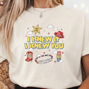 May include: A cream-colored t-shirt featuring a graphic print with the text "I KNEW IT I KNEW YOU" in yellow with blue outlines. The design includes a gold sheriff badge, a red cowboy hat, red cowboy boots, a beaded bracelet, and alphabet blocks.