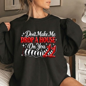 May include: A black crewneck sweatshirt featuring the white and red text "Don't Make Me DROP A HOUSE On You" above a graphic of two legs wearing black and white striped stockings and shiny red high-heeled shoes, accented with small white stars.
