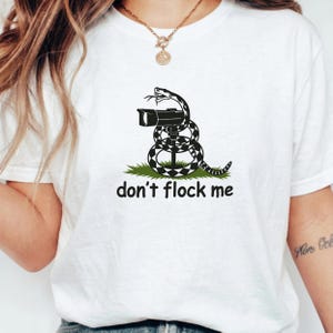 Puede incluir: Una camiseta blanca con un gráfico en blanco y negro de una serpiente enroscada alrededor de un objetivo de cámara, sobre un parche de hierba verde. Debajo del gráfico, el texto "don't flock me" está impreso en una fuente negra y negrita.