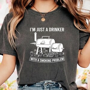 Può includere: Una t-shirt grigio antracite con una grafica bianca di un barbecue smoker circondato da lattine di bevande sparse. Il testo I'M JUST A DRINKER WITH A SMOKING PROBLEM è stampato in un carattere bianco, grassetto e senza grazie sul petto.