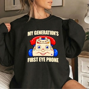 Puede incluir: Una sudadera negra de manga larga con el gráfico de un teléfono de juguete vintage con auricular rojo, ruedas azules y una cara sonriente. El texto superior dice MY GENERATION'S y el texto inferior dice FIRST EYE PHONE en letras blancas.