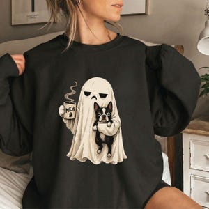 May include: A black crewneck sweatshirt featuring a graphic of a white ghost with a grumpy expression holding a steaming mug labeled "MEH" and carrying a black and white Boston Terrier dog. The design is centered on the front of the garment.