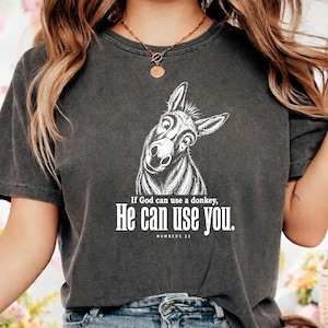 Puede incluir: Una camiseta gris carbón de manga corta con una ilustración de líneas blancas de la cabeza de un burro. Debajo del gráfico, el texto dice: If God can use a donkey, He can use you. NUMBERS 22. La camiseta tiene cuello redondo y un acabado desgastado.