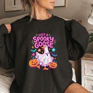 Könnte beinhalten: Ein schwarzes Langarm-Sweatshirt mit einem Grafikdruck einer Gans in einem weißen Geisterkostüm. Das Design umfasst zwei orangefarbene Kürbisse, violette Fledermäuse sowie blaue und gelbe Sterne mit dem Text JUST A SPOOKY GOOSE in Pink.