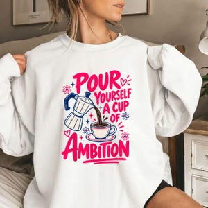 Op de afbeelding: Een wit sweatshirt met lange mouwen en een grafische print van een mokkapot die koffie in een kopje schenkt. De tekst "POUR YOURSELF A CUP OF AMBITION" staat in dikke roze en blauwe letters, versierd met kleine roze hartjes, sterretjes en bloemen.