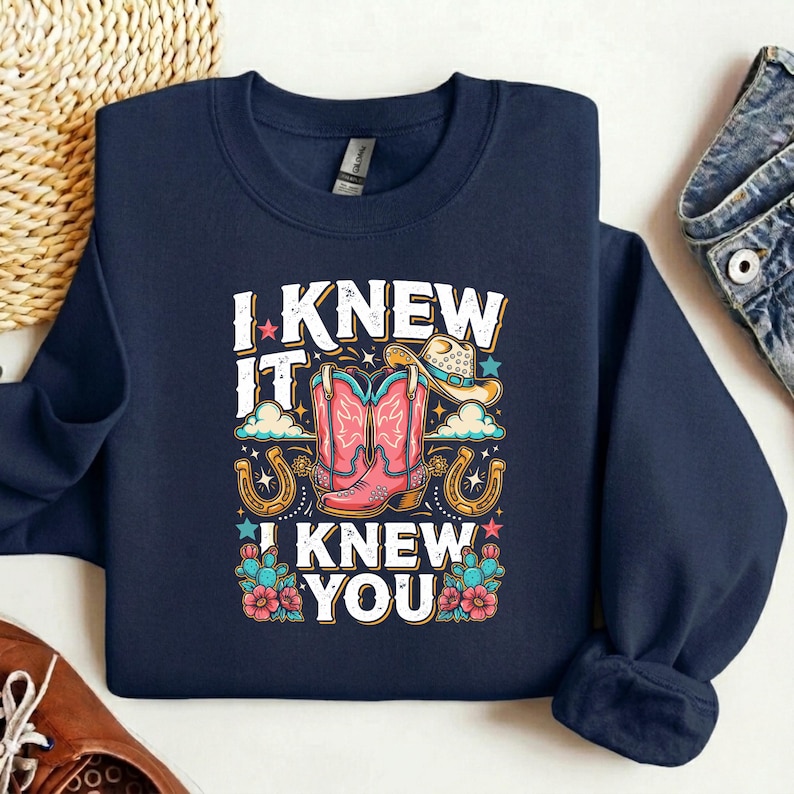 Op de afbeelding: Een marineblauw sweatshirt met ronde hals en een grafische print met de tekst I KNEW IT I KNEW YOU. Het ontwerp bevat roze cowboylaarzen, een beige cowboyhoed, twee gouden hoefijzers, wolken, sterren en kleurrijke woestijnbloemen en cactussen.