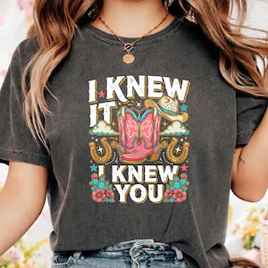 Op de afbeelding: Een antracietgrijs T-shirt met korte mouwen en een grafische print met de tekst I KNEW IT I KNEW YOU, roze cowboylaarzen, een beige cowboyhoed, gouden hoefijzers, blauwe cactussen met roze bloemen en kleine kleurrijke sterren en wolken.