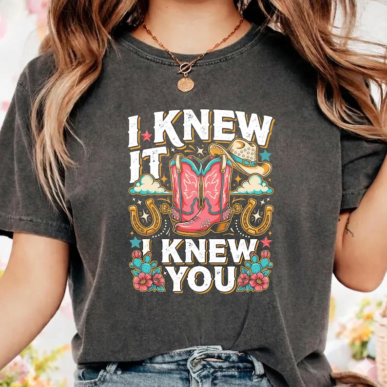 Op de afbeelding: Een antracietgrijs T-shirt met korte mouwen en een grafische print met de tekst I KNEW IT I KNEW YOU, roze cowboylaarzen, een beige cowboyhoed, gouden hoefijzers, blauwe cactussen met roze bloemen en kleine kleurrijke sterren en wolken.