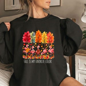 May include: A black long-sleeve sweatshirt featuring a graphic of five pink flamingos standing among autumn trees with red, orange, and yellow leaves. The flamingos wear scarves and hats. The text below reads, Fall Is My Favorite Color.